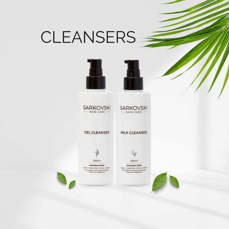 Two bottles of SARKOVSKI® Skin Care Milk Cleanser and Gel Cleanser displayed on a marble surface with natural elements like leaves, a white rock, and a piece of bark, emphasizing their clean and natural formulation.
