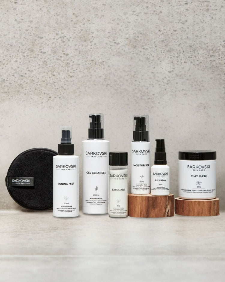 A set of SARKOVSKI® Skincare Gel Cleanser, including a toning mist, gel cleanser, exfoliant, moisturiser, eye cream, and clay mask, elegantly arranged on wooden risers against a textured background.