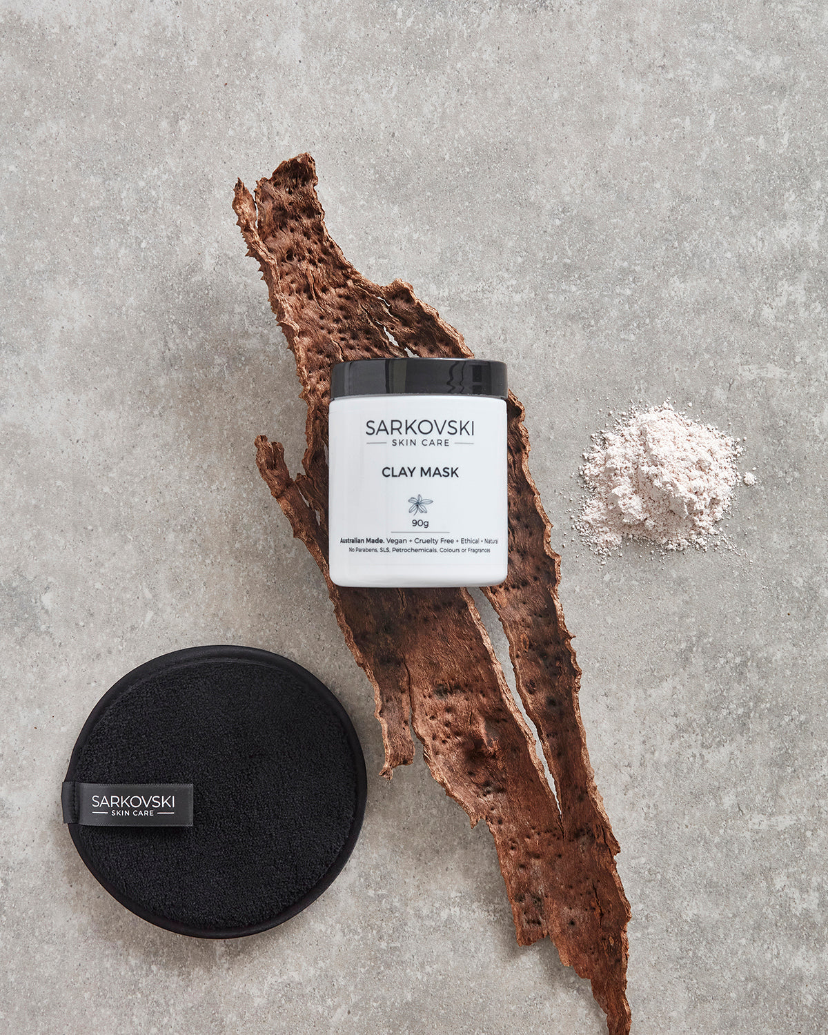 SARKOVSKI® Clay Mask on textured stone with bark, clay powder, and black cleansing cloth, emphasizing natural, earthy elements in skincare.
