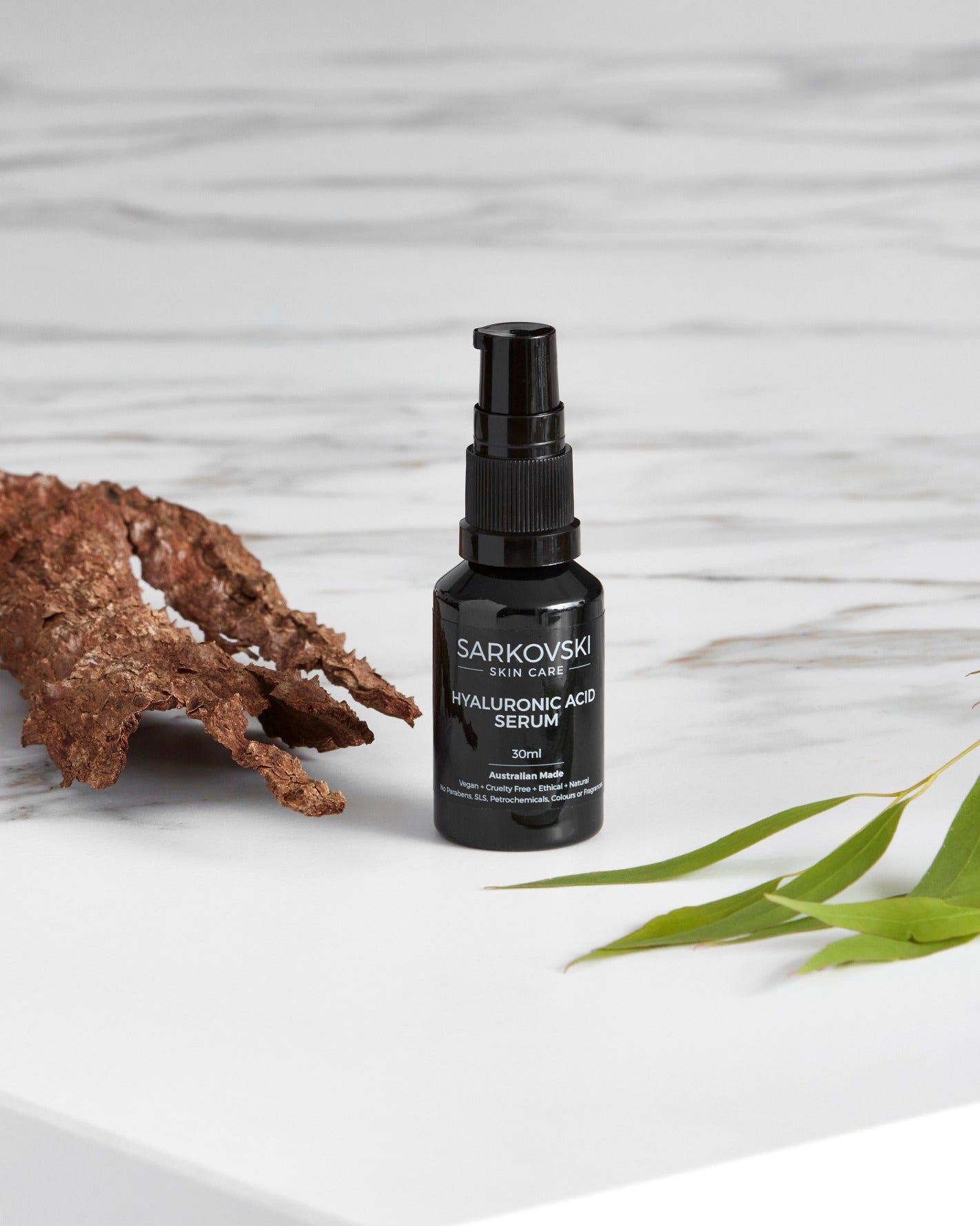 SARKOVSKI Hyaluronic Acid Serum bottle displayed on a marble surface beside natural elements like bark and green leaves, showcasing a luxurious and nature-inspired skincare aesthetic.