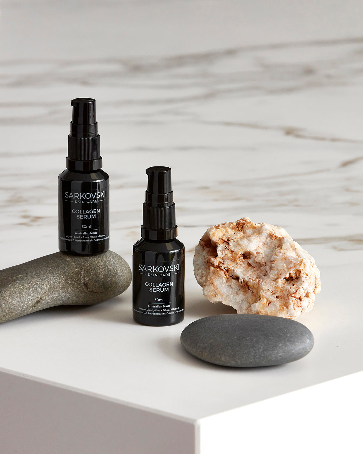 SARKOVSKI® Natural Skin Care Collagen Serum - two bottles of Collagen Serum with two smooth stones. One Collagen Serum bottle on top of one stone. All are neatly arranged on top of marble table. Collagen Serum is designed to boost collagen, hydrate deeply, and restore skin elasticity.