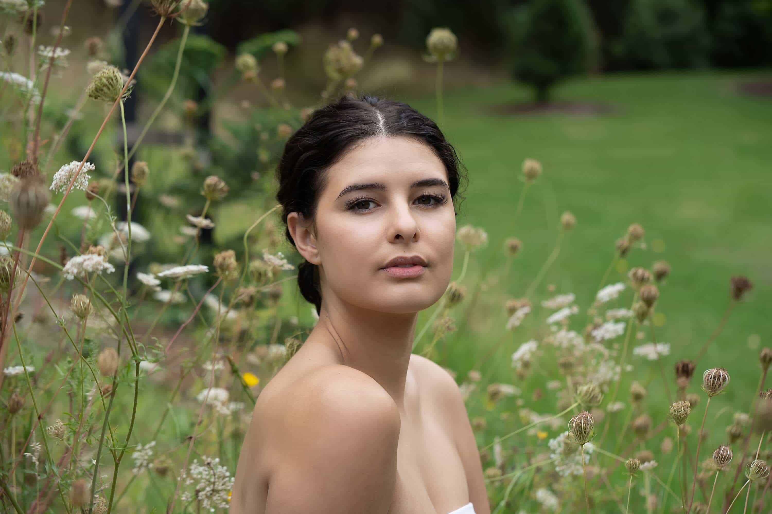 A young woman with dark curly hair posing in a field of wildflowers, gazing softly at the camera with a serene expression.