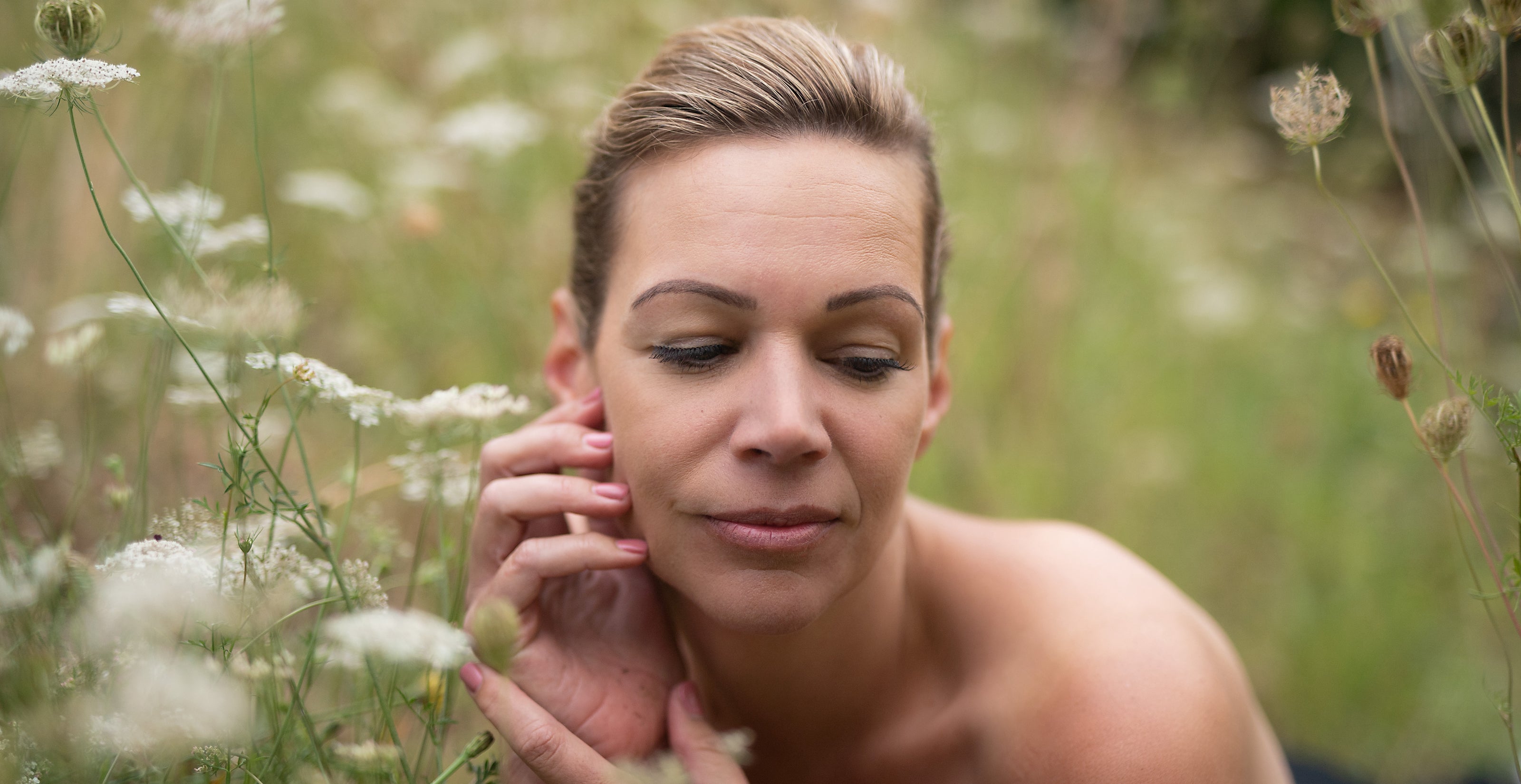 A woman with blonde hair touching her face in a field of wildflowers, gazing into the distance with a serene expression.
