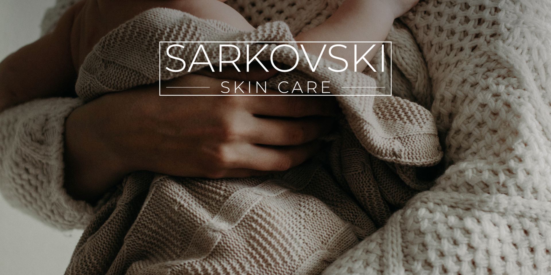 A close-up of a mother gently cradling her baby, both wrapped in soft, beige knitwear. The image features the SARKOVSKI Skin Care logo overlay, suggesting comfort, tenderness, and natural skincare during pregnancy and breastfeeding.
