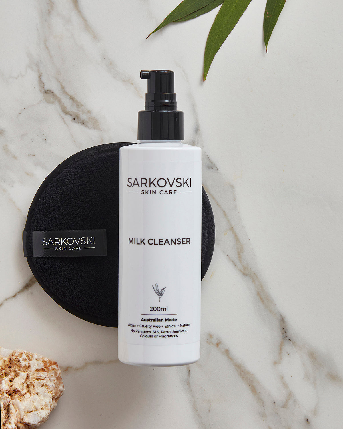 A sleek white bottle of SARKOVSKI® Skin Care Milk Cleanser with a black pump, placed on a marble surface alongside the SARKOVSKI® Makeup Removal Cleansing Cloth. The setting includes green leaves and a textured stone, highlighting a natural and luxurious skincare theme.