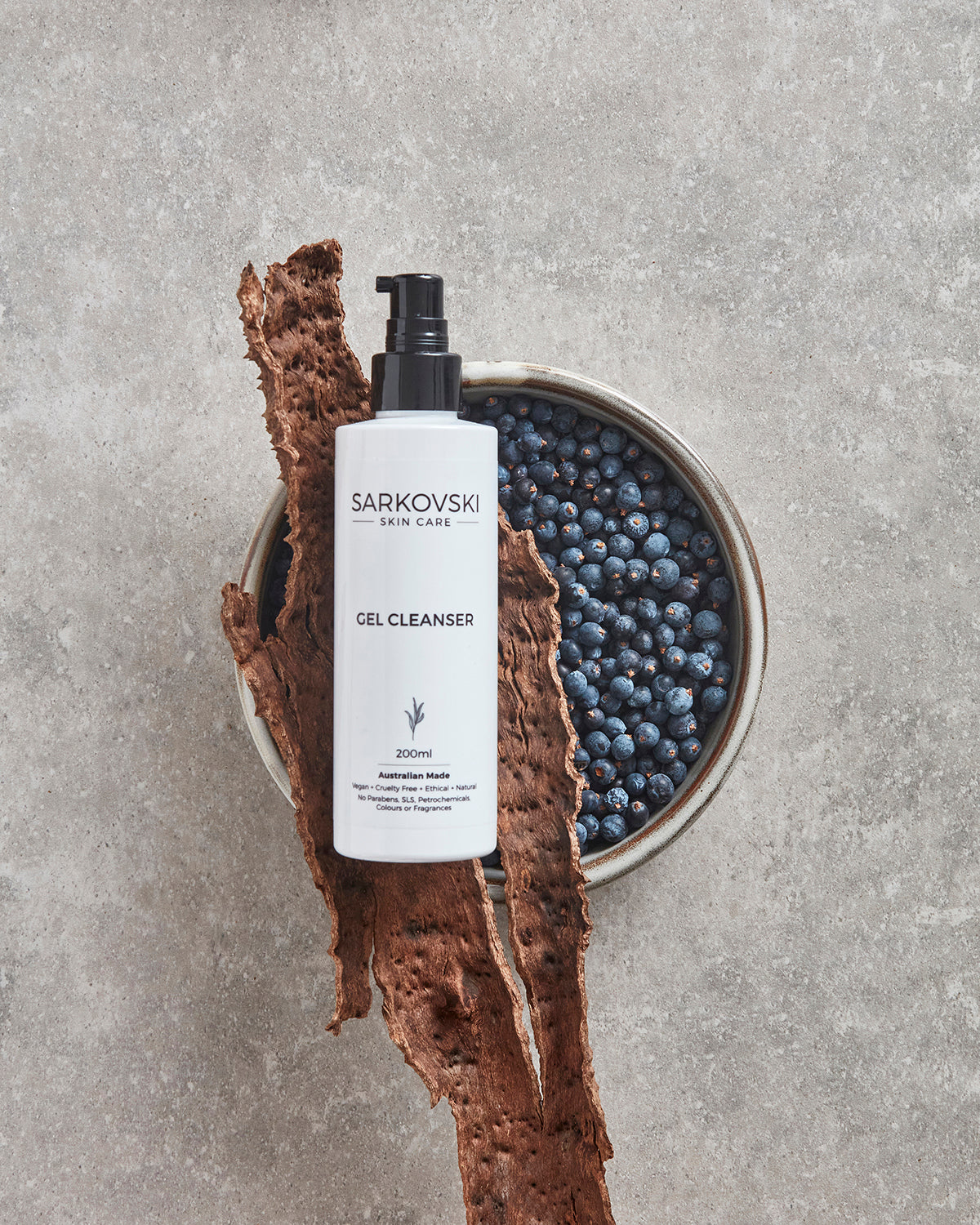 SARKOVSKI Gel Cleanser bottle on a cemented surface, surrounded by dark berries and rustic bark, emphasizing its natural ingredients and hydrating formula.